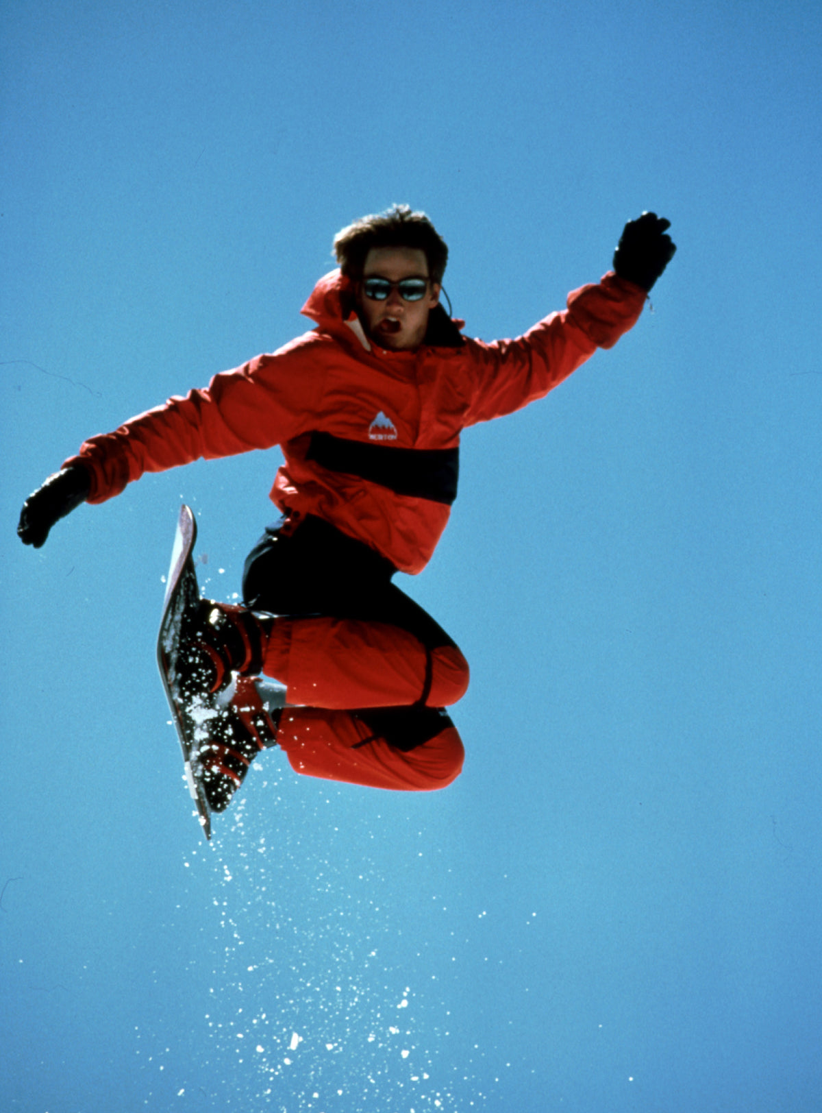 A snowboarder in a bright red and black ski suit jumps high against a clear blue sky, with snow spraying from the board.