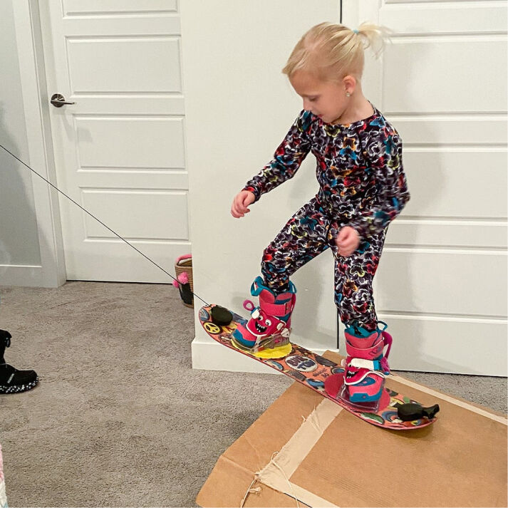 A child riding a Riglet snowboard indoors on carpet with a cardboard jump ramp
