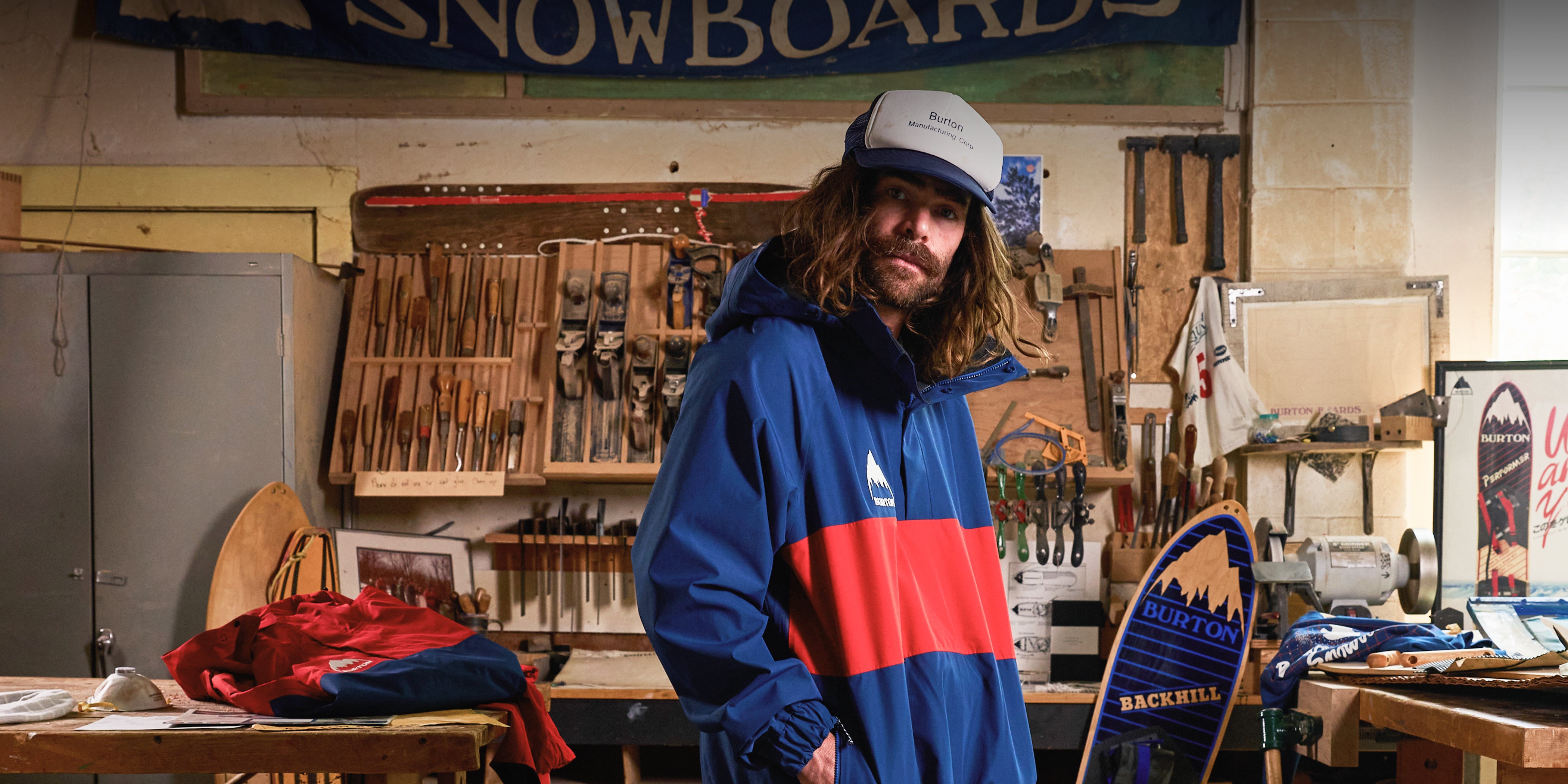 Danny Davis wearing a blue and red snowboard jacket and a trucker cap, standing in a workshop filled with snowboard equipment and tools.