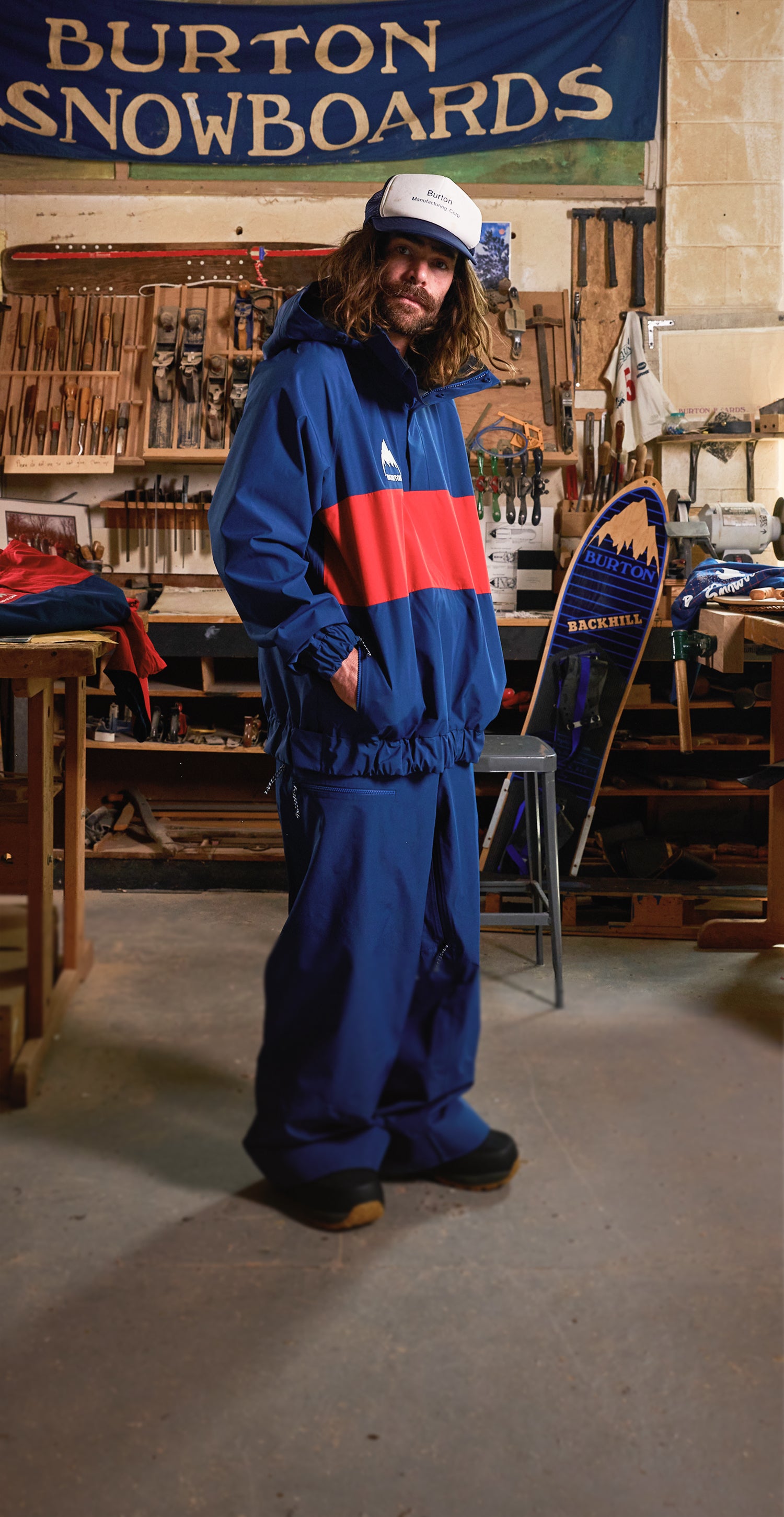 Danny Davis wearing a blue and red snowboard jacket and a trucker cap, standing in a workshop filled with snowboard equipment and tools.