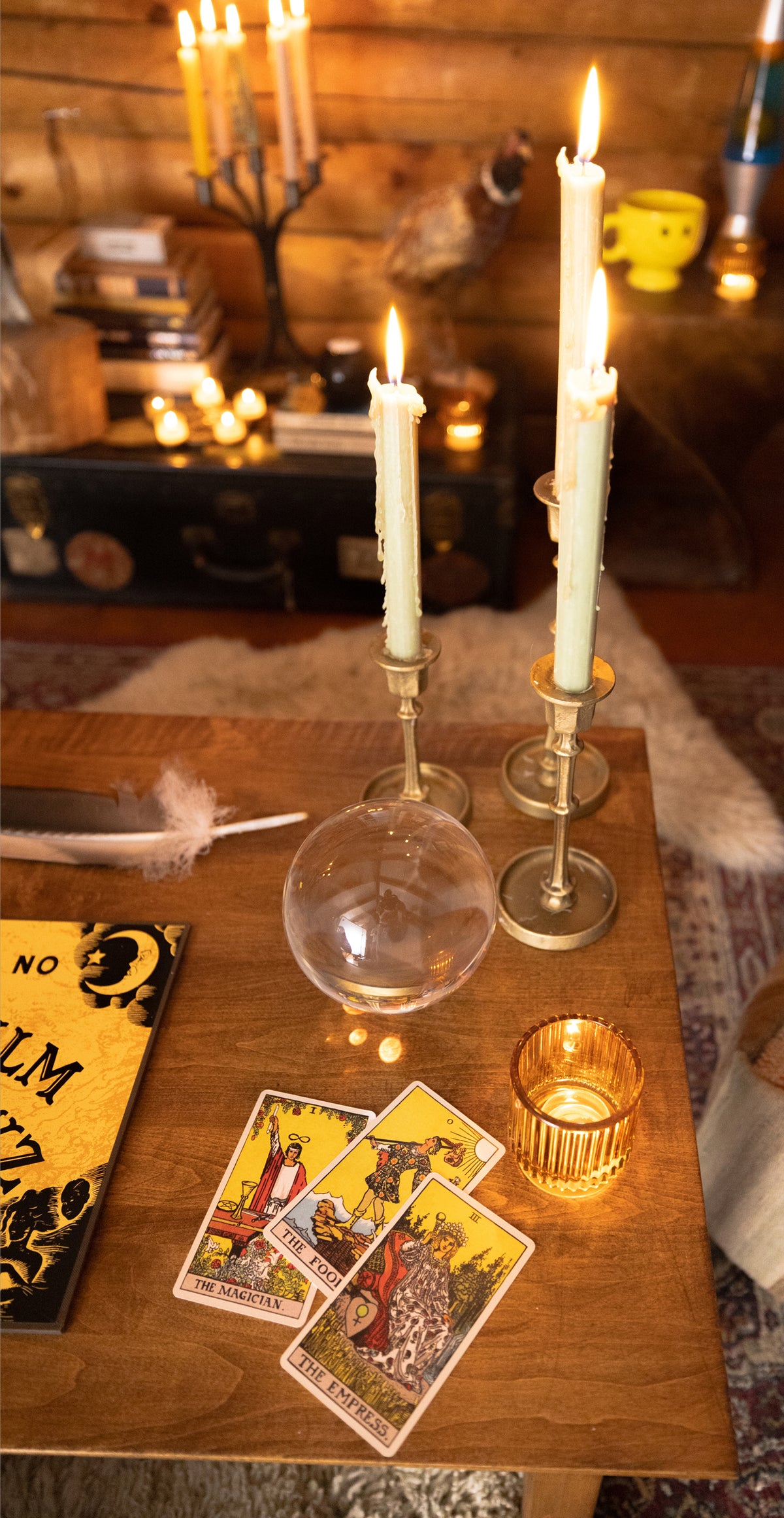 A cozy tabletop scene with lit white candles in brass holders, a clear glass orb, a small amber candle, tarot cards, and a yellow book on a wooden table in a warmly lit room.