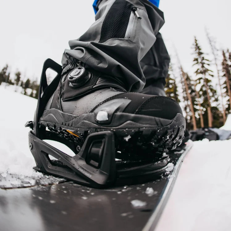 Close-up of a snowboard boot attached to a snowboard with a snowy landscape in the background.