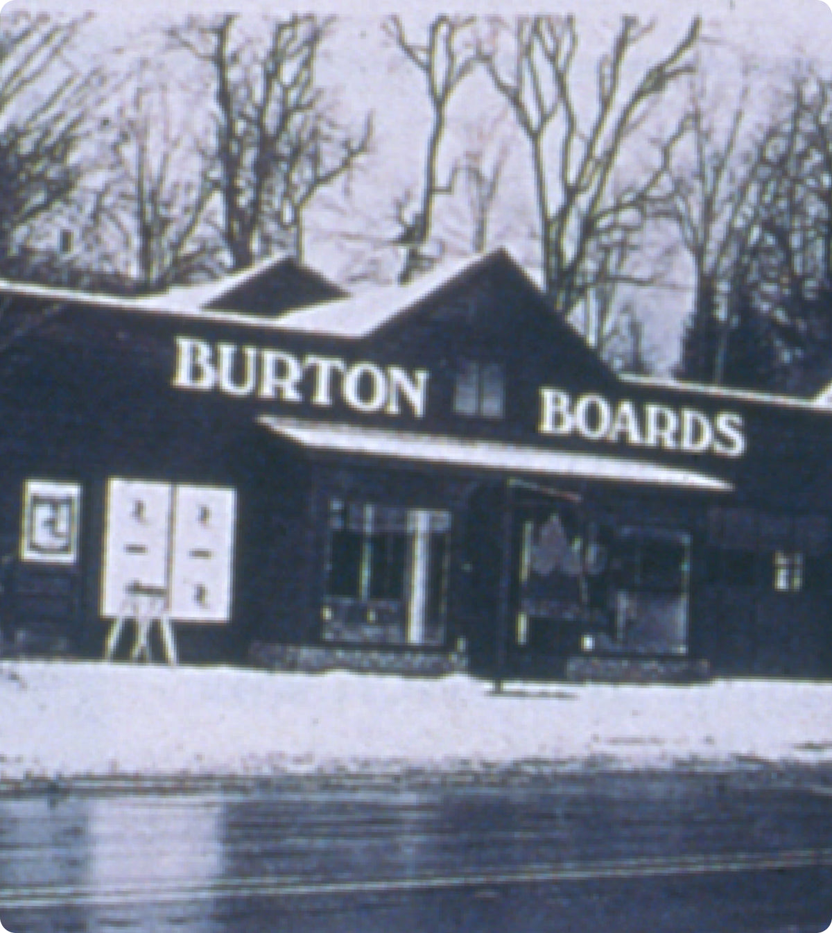 A vintage black-and-white photograph of the Burton Snowboards storefront in Manchester, VT, with snow on the ground and bare trees in the background. Large white text on the building reads BURTON BOARDS.