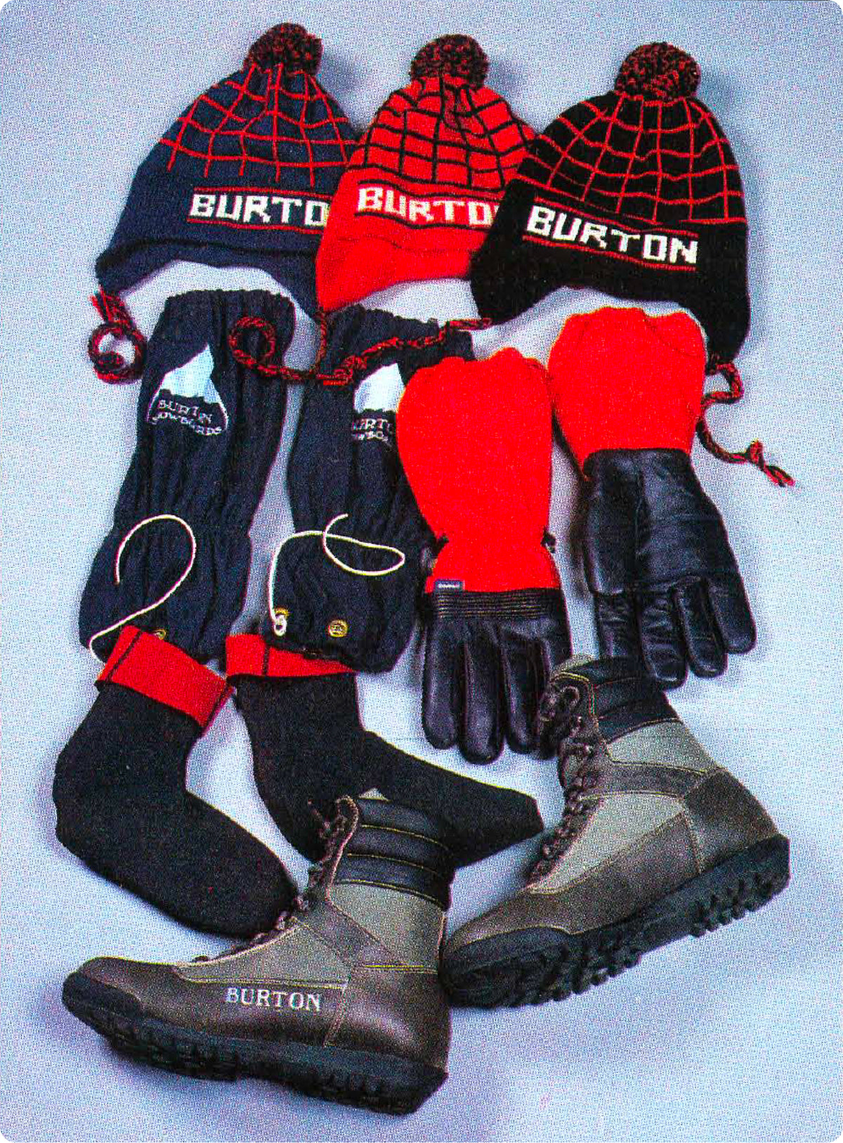 A flat lay of vintage Burton winter gear from the early 1980s including three black and red web-pattern knit beanies with white BURTON logos, matching knit gloves, black socks, and brown and gray boots on a light gray background.