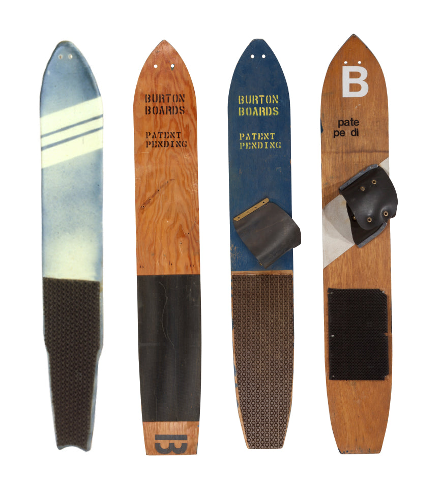 Four vintage Burton snowboards displayed upright against a white background, featuring different wood grain finishes, black grip panels, and printed branding.