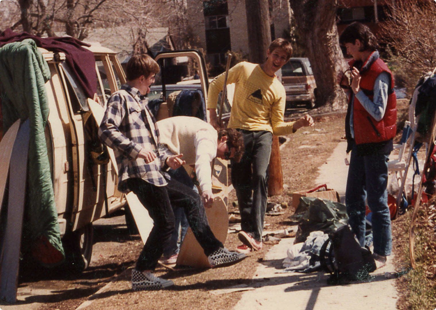 A group of four young men standing outdoors on a sidewalk next to parked vans and trees, wearing casual 1980s streetwear. One man in a yellow long-sleeve shirt is prominent, and a small inset in the bottom right corner shows a black long-sleeve shirt with a small chest logo.