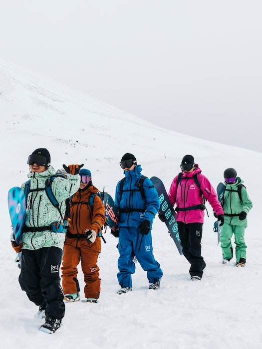 Five people in colorful winter gear walking on a snow-covered slope with snowboards.