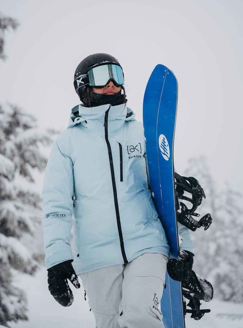 A woman in a light blue jacket carrying a splitboard and walking through snowy forest