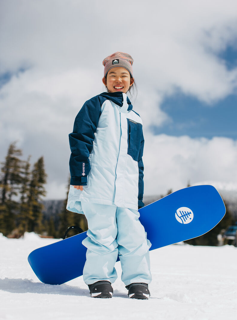 Child standing facing front holding a snowboarding and dressed in a snow jacket and snow pants